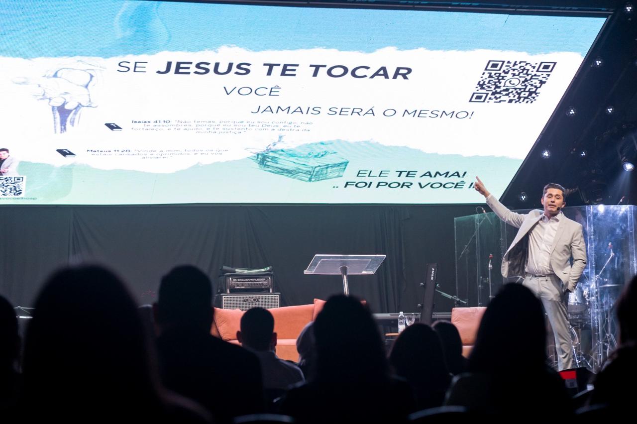 Gustavo Coelho palestrando em grande auditório, apontando para tela com a mensagem Se Jesus te tocar